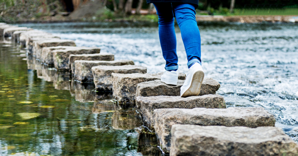 person in jeans and white shoes stepping across stones to cross the river